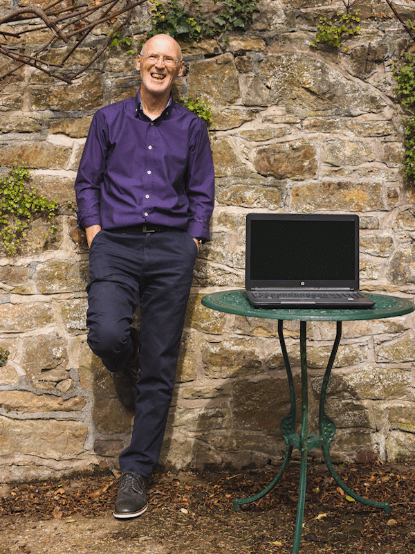 Eric Davies of DoodlyDog website design in purple shirt standing against a wall next to laptop computer on a green metal table.